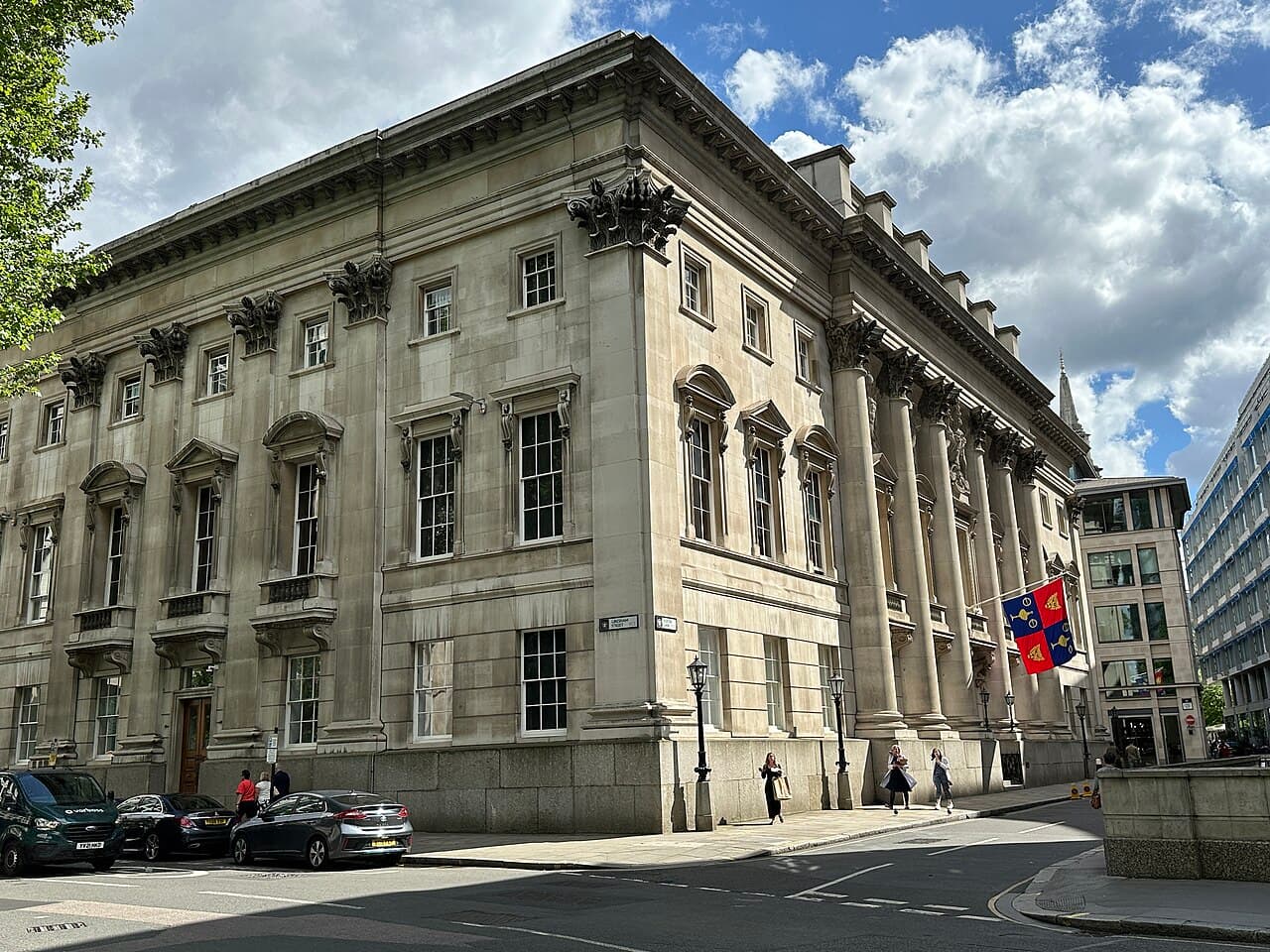 A large pale stone building with tall columns on a London street corner, flying a red, blue and gold flag.