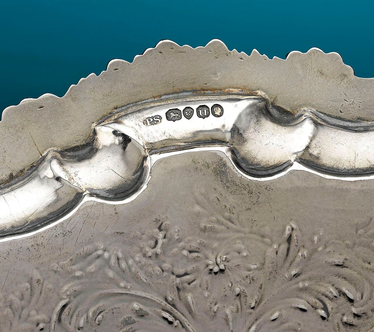 Close-up of the rim of a silver tray stamped with a row of five small marks: the initials PS, a walking lion, a leopard's face, a letter u and a man's head in profile.