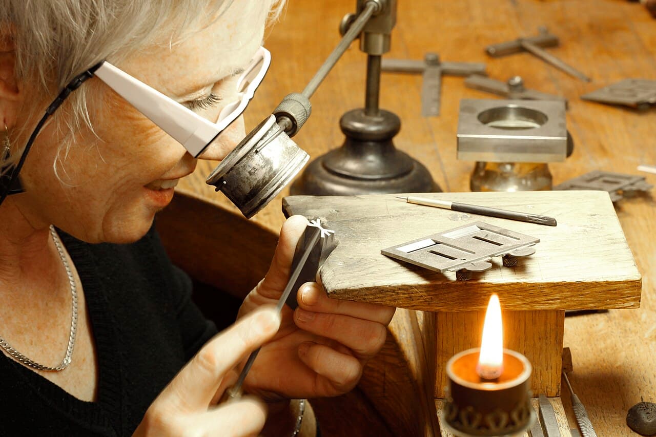 A white-haired woman wearing magnifying glasses holds a small steel punch against a worn wooden bench; a small flame burns in a holder in the foreground.