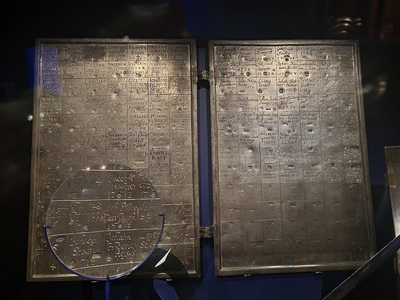 Two hinged grey metal plates in a dark museum case, divided into a grid; each cell holds an engraved name, a year and small struck marks, with a magnifying lens in front.